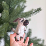 A handmade wool felt pug finger puppet is held by a female hand against a festive Christmas tree. This charming Christmas felt finger puppet features a tan body, brown markings on its face, and floppy brown ears, with visible feet and a tail.