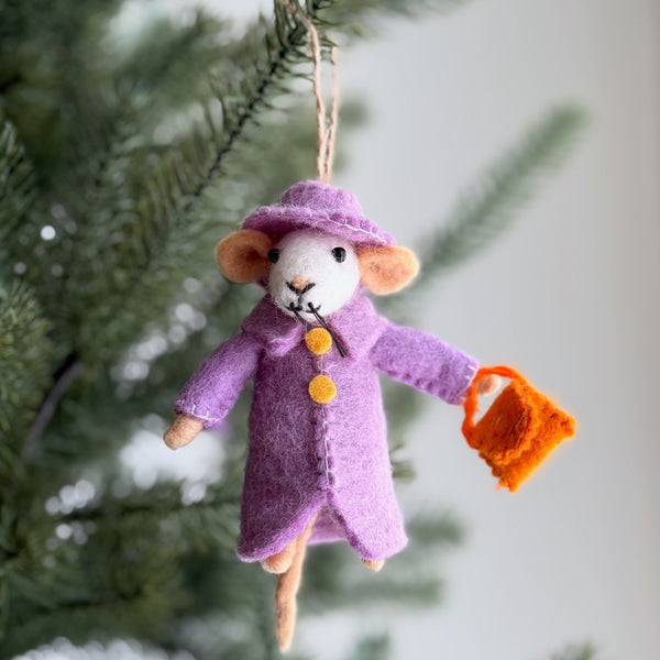 The Queen Mousiebeth wool felt mouse ornament hangs from a Christmas tree branch by a jute string, against a softly blurred festive background. This charming handcrafted felt ornament features the mouse wearing a regal lavender coat and matching wide-brimmed hat, holding an orange felt handbag, with delicate needle-felted details.