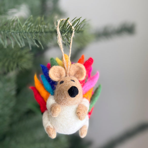 A cheerful wool felt rainbow hedgehog ornament hangs on a Christmas tree. This handmade felt ornament features a light tan, rounded hedgehog body with vibrant felt spikes in blue, green, yellow, orange, pink, and red. It has small ears, a black nose, and a neutral expression, completed with a jute string for easy hanging.