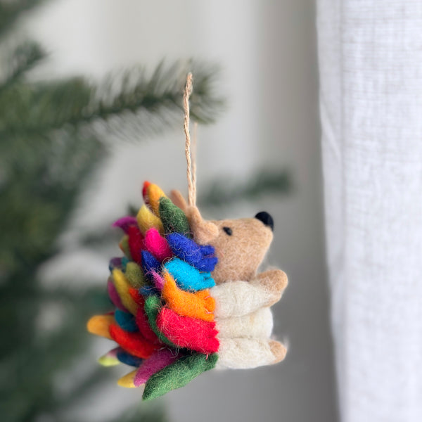 A vibrant rainbow hedgehog ornament is suspended on a Christmas tree, set against a soft, blurred background. This wool felt ornament features bright rainbow-colored spikes, transitioning from green, red, orange, yellow, blue, and magenta, with a tan face and body, tiny black nose, and ears. It hangs by a rustic jute string, adding a joyful, festive touch.