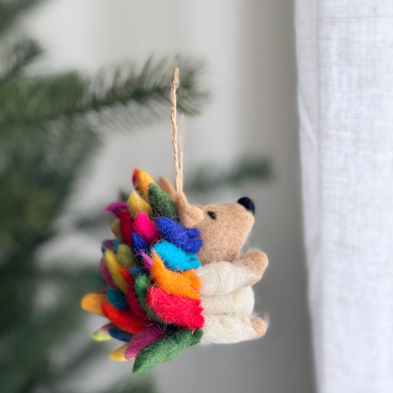 A vibrant rainbow hedgehog ornament is suspended on a Christmas tree, set against a soft, blurred background. This wool felt ornament features bright rainbow-colored spikes, transitioning from green, red, orange, yellow, blue, and magenta, with a tan face and body, tiny black nose, and ears. It hangs by a rustic jute string, adding a joyful, festive touch.