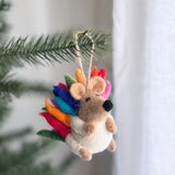 A wool felt rainbow hedgehog ornament hangs from a vibrant green Christmas tree branch, creating a joyful and festive atmosphere. This charming Christmas tree ornament features rainbow-colored spikes across its back, transitioning through red, orange, yellow, green, blue, and pink. It has a light brown face with small ears, a black nose, and folded arms over a fluffy white belly. A jute string allows the unique Christmas ornament to dangle gracefully.