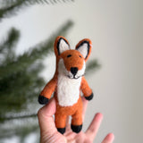 A handcrafted wool felt Red Fox finger puppet is gently held by a female hand, set against a soft, diffused background hinting at a Christmas tree. This unique felt fox puppet features a vibrant red hue with black accents on its paws and ear outlines, showcasing its detailed craftsmanship.