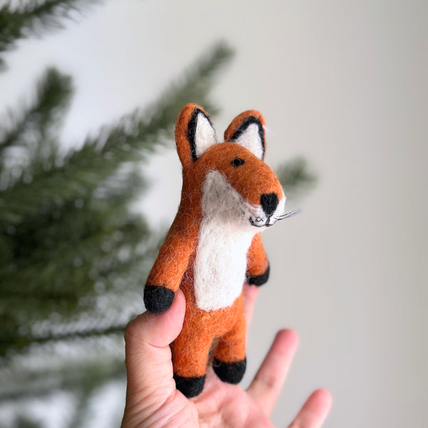 A handmade red fox wool felt finger puppet is gently presented by a female hand against a soft white backdrop with a hint of a Christmas tree. This charming, meticulously crafted felt fox puppet features a white belly, inner ears, and muzzle. Black accents on its feet, nose, and ear tips, along with delicate whiskers, complete its endearing design.
