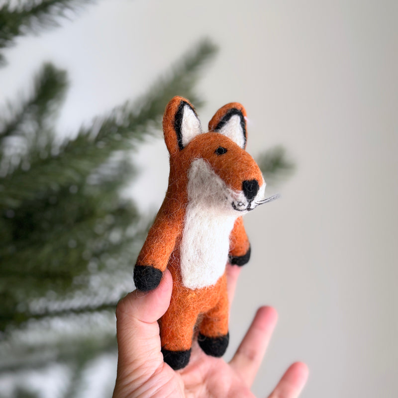 A handmade red fox wool felt finger puppet is gently presented by a female hand against a soft white backdrop with a hint of a Christmas tree. This charming, meticulously crafted felt fox puppet features a white belly, inner ears, and muzzle. Black accents on its feet, nose, and ear tips, along with delicate whiskers, complete its endearing design.