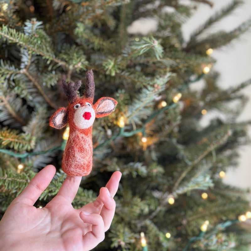 A festive needle felted Red Reindeer Finger Puppet rests on a female hand, with a blurred Christmas tree and yellow lights in the background creating a warm holiday setting. This charming felt reindeer puppet is primarily red and cream, featuring brown antlers and a distinct red nose.