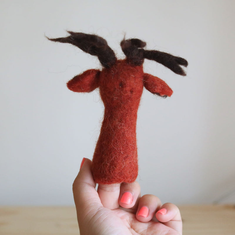 A red reindeer wool felt finger puppet is held upright by a female hand against a plain, light background. This charming handmade felt toy for kids features a vibrant red body, a cute red nose, and darker brown antlers. The hand, with coral nail polish, showcases the puppet's soft texture and unique craftsmanship.