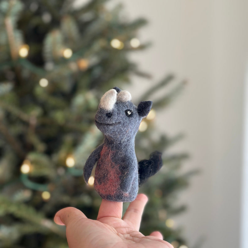 A grey needle-felted rhino finger puppet is held by a hand, set against a blurred Christmas tree with soft, warm lights, creating a playful, whimsical scene. This handcrafted felt puppet features a charming design with a soft, fuzzy texture, and includes a distinctive white horn, small arms, and a tail.