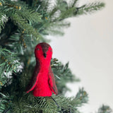 A handcrafted felt robin finger puppet is perched on an indoor Christmas tree branch, set against a bright background. This unique, ethically made robin puppet has a round red and brown body, a dark brown head, and small dark wings, reflecting the fair-trade craftsmanship from Nepal.