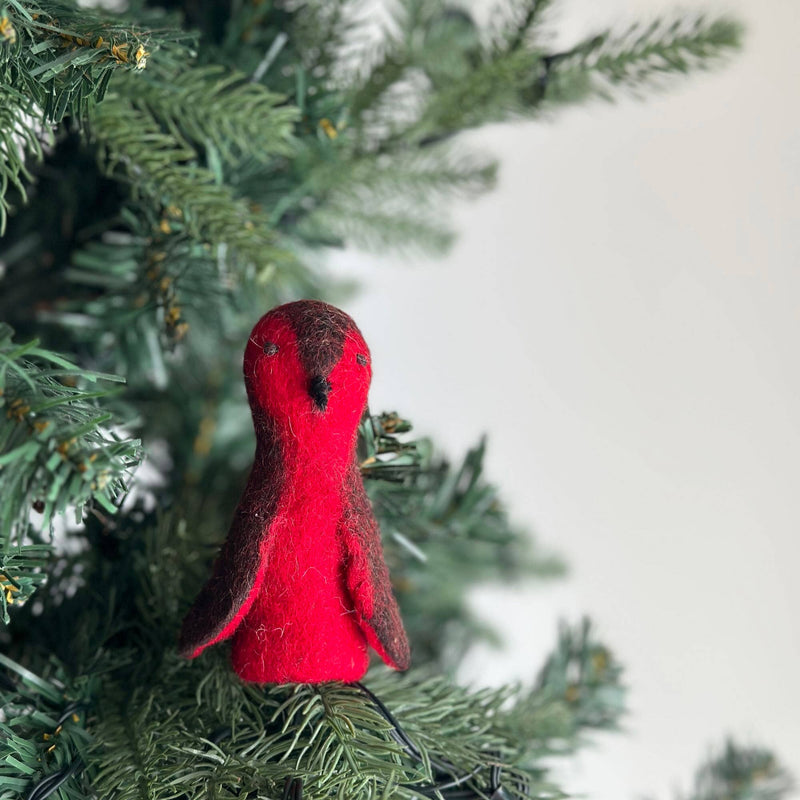 A handcrafted felt robin finger puppet is perched on an indoor Christmas tree branch, set against a bright background. This unique, ethically made robin puppet has a round red and brown body, a dark brown head, and small dark wings, reflecting the fair-trade craftsmanship from Nepal.