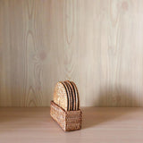 A detailed close-up of a handcrafted Bali rattan coaster set of 5, handmade in Bali. The five round coasters, featuring a tight spiral weave and thick braided edges, are stacked vertically inside a matching rectangular woven rattan holder on a light wood surface.
