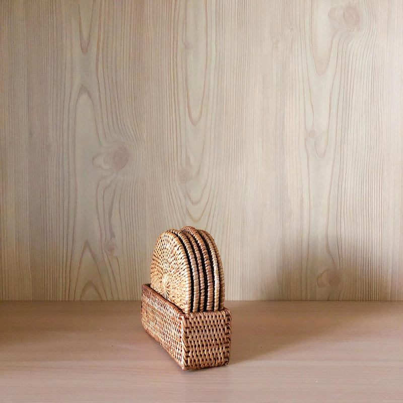 A detailed close-up of a handcrafted Bali rattan coaster set of 5, handmade in Bali. The five round coasters, featuring a tight spiral weave and thick braided edges, are stacked vertically inside a matching rectangular woven rattan holder on a light wood surface.