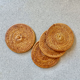 Handcrafted Bali rattan coaster set of 5, handmade in Bali, seen from a top-down perspective against a cool-toned, grey speckled surface. Four golden-brown coasters are artfully arranged; one lies flat on the left, showcasing its intricate sunburst weave, while three others overlap in a casual stack on the right. The warm, natural texture of the Bali rattan coaster set creates a beautiful organic contrast with the sleek, modern background, evoking a rustic yet sophisticated feeling.