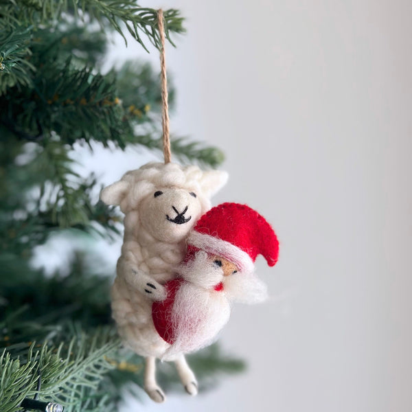 A charming Santa's Little Helper wool felted sheep ornament hangs gracefully on a Christmas tree, attached by a jute string. The snowy white sheep features small, stitched eyes and a smiling mouth. It cradles a miniature Santa doll with a red hat and white beard in its tiny hooves, adding handcrafted charm to the holiday decor.