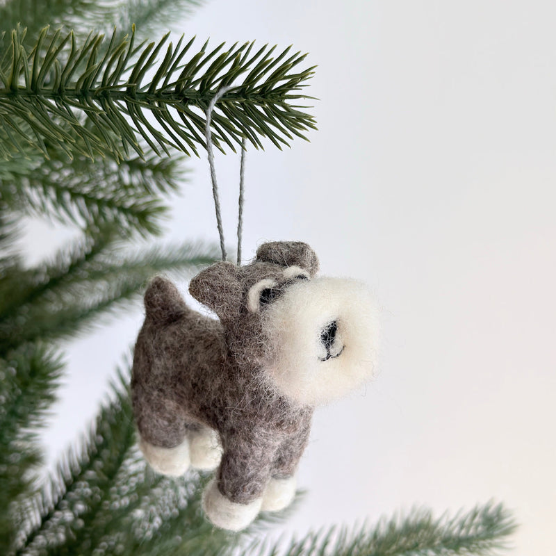 A felt Schnauzer dog ornament hangs from a vibrant green Christmas tree branch. The gray Schnauzer dog features a fluffy white beard and eyebrows, white paws, and a cute smile, suspended by a gray string.