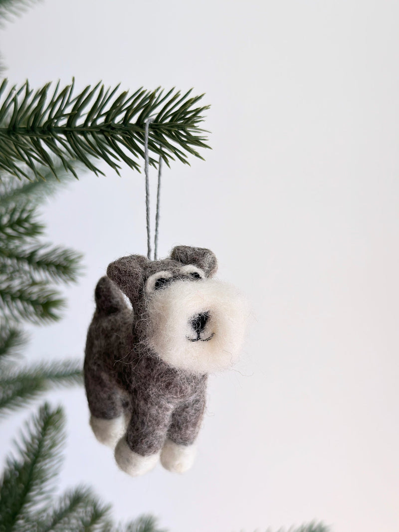 A felt Schnauzer dog ornament hangs from a vibrant green Christmas tree branch against a white backdrop. The felt Schnauzer ornament has a fluffy white beard, fluffy eyebrows, and a black stitched smile. Its body is grey with white paws, and a grey cotton string is attached for hanging. This unique Christmas tree ornament evokes warmth and festive cheer.