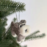 A gray and white needle felted Schnauzer dog ornament is suspended from a festive Christmas tree branch. This adorable handcrafted felt ornament features a fluffy beard, prominent eyebrows, and a cute smile, ready to add charm to your holiday decor.