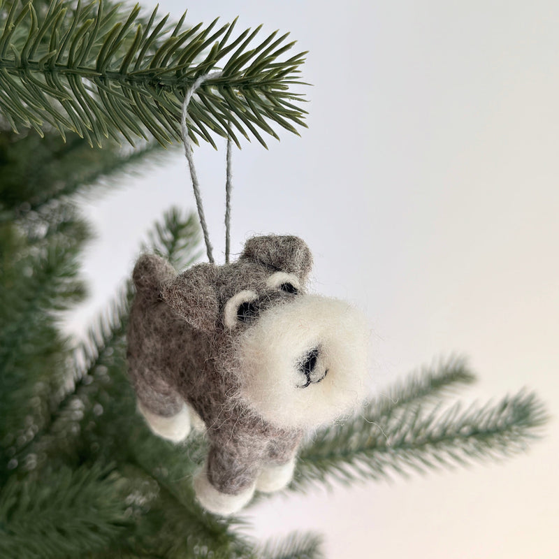 A gray and white needle felted Schnauzer dog ornament is suspended from a festive Christmas tree branch. This adorable handcrafted felt ornament features a fluffy beard, prominent eyebrows, and a cute smile, ready to add charm to your holiday decor.