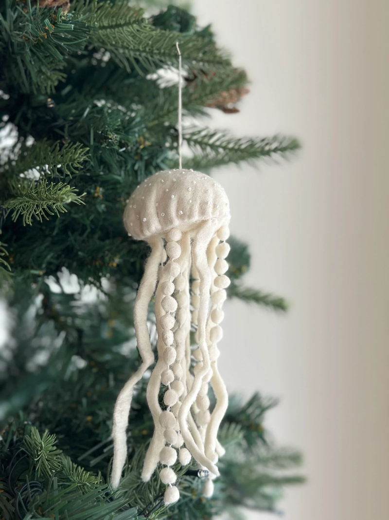 An off-white felt jellyfish, part of a Sea Life Garland baby mobile, hangs on a dark green Christmas tree against an off-white wall. Its round, bell-shaped body is adorned with small white embellishments. Long, thick felt tentacles extend downwards, some featuring small felt balls at even intervals.