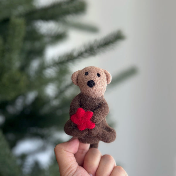 A female hand holds a handcrafted needle felted sea otter finger puppet, featuring a vibrant red felt sea star, against a blurred Christmas tree backdrop. The charming wool felt sea otter puppet has a brown body, lighter brown face, small rounded ears, and black eyes. Its artisan design offers a warm, inviting feel.