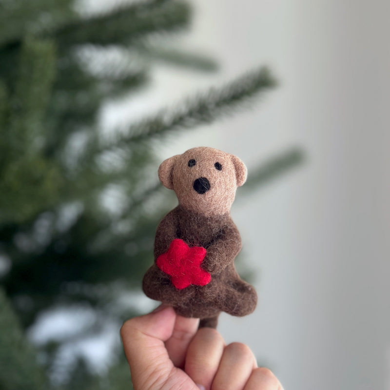 A female hand holds a handcrafted needle felted sea otter finger puppet, featuring a vibrant red felt sea star, against a blurred Christmas tree backdrop. The charming wool felt sea otter puppet has a brown body, lighter brown face, small rounded ears, and black eyes. Its artisan design offers a warm, inviting feel.