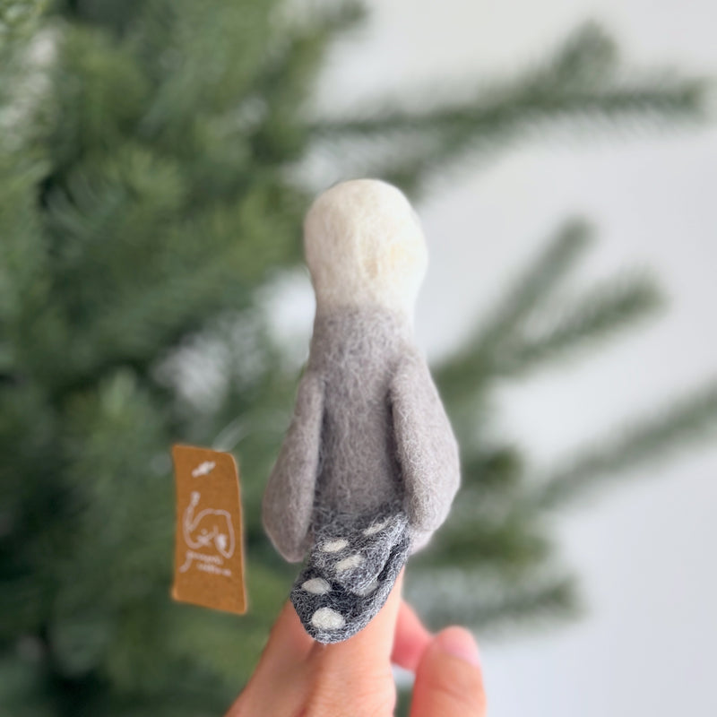 A handmade felt seagull finger puppet, worn on a female hand, is set against a blurred Christmas pine tree background. This unique seagull puppet features a white head, a light gray body, and a darker gray tail with white dots, embodying fine craftsmanship.