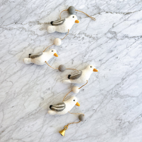 A charming wool felt seagull garland is displayed on a marble-patterned surface, creating a calm and playful atmosphere. It features four white felt seagulls with grey wing accents and small yellow beaks, evenly spaced along a natural fiber string. Alternating grey and white wool balls separate each seagull, and a small copper bell hangs at the bottom.