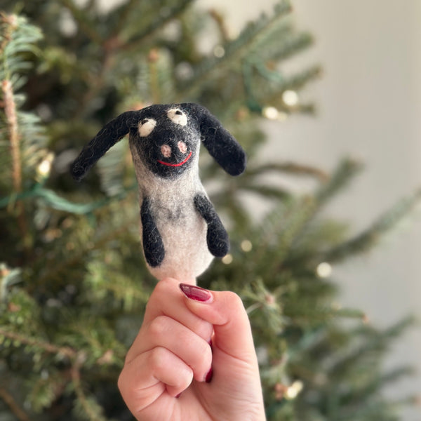 A handmade wool felt Shaun the Sheep finger puppet is held by a female hand with dark red nail polish, positioned against a blurred Christmas tree background. This unique needle felted puppet, crafted from 100% wool, features distinctive black ears, a pink snout, and a small red smile, creating a playful and festive mood.