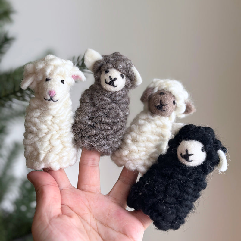 A handmade felt sheep finger puppet is presented on a female hand against a blurred backdrop of green foliage, evoking a playful and cozy atmosphere. This unique felted finger puppet features fluffy, textured wool, small ears, and a simple embroidered face, adding to its whimsical charm and inviting creative play.