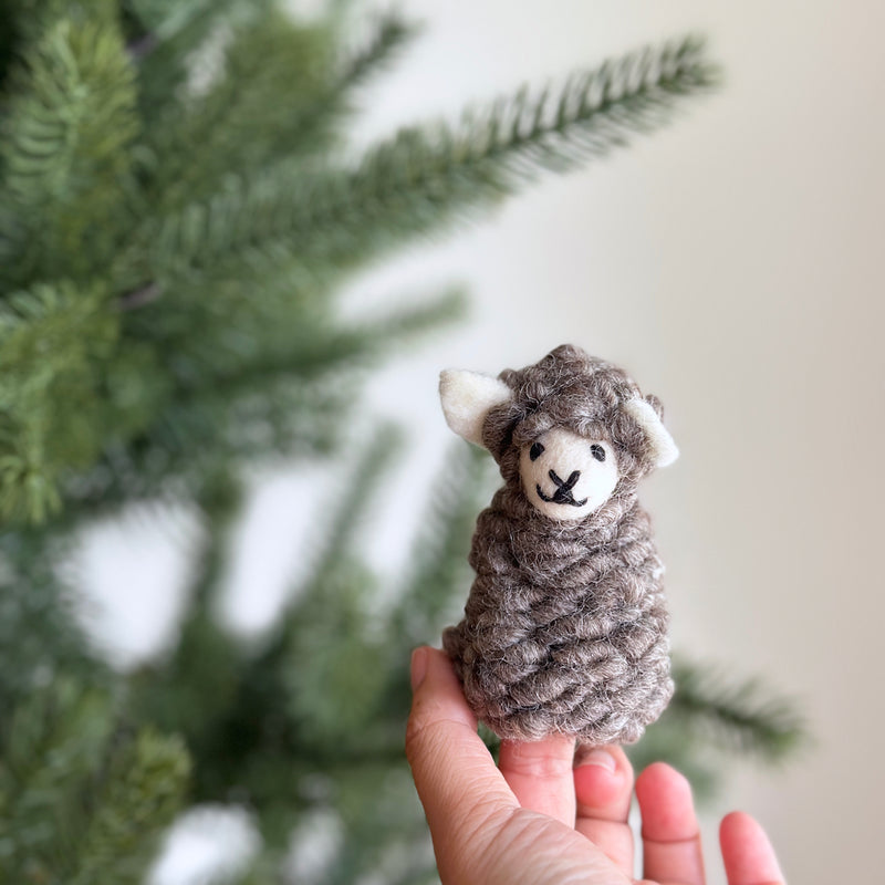 A charming handcrafted felt sheep finger puppet is held by a female hand, set against a soft, neutral backdrop with a blurred green pine tree. This unique felt finger puppet has a fluffy, textured body in muted brown tones and small, light beige ears. Its sweet face features stitched black eyes and an 'X' mouth, giving it a friendly appearance.