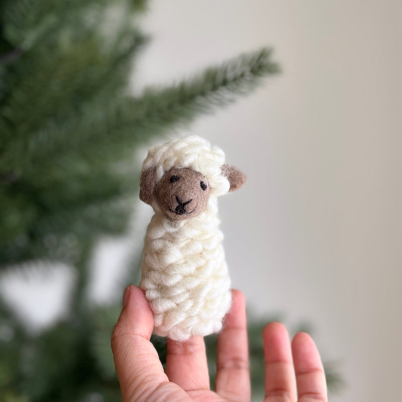 A handmade wool felt sheep finger puppet is gently presented by a hand, set against a soft, blurred indoor background with subtle green foliage. This endearing, cream-colored sheep features a fluffy wool felt body, brown face and ears, and a stitched black 'X' for its nose. Its round shape and textured design make it a charming felt toy for imaginative play.