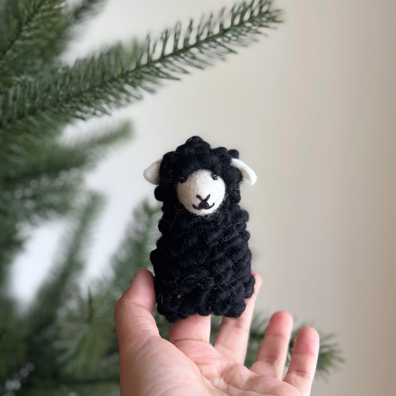 A felt sheep finger puppet is held in a female hand, set against a blurred green pine tree branch and a light beige wall. This sheep puppet features a fluffy black body and a white face with small white ears. Its detailed and fluffy texture creates a cuddly and playful appearance, enhancing the soft, natural atmosphere.