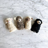 Several handmade felt sheep finger puppets are arranged in a row on a textured white marble surface. These cuddly felt sheep toys showcase a soft, fluffy texture and charming 3D designs. Visible puppets include a white sheep with pink inner ears, a brown sheep, a cream-colored sheep with a brown face, and a black sheep, each with a smiling face.