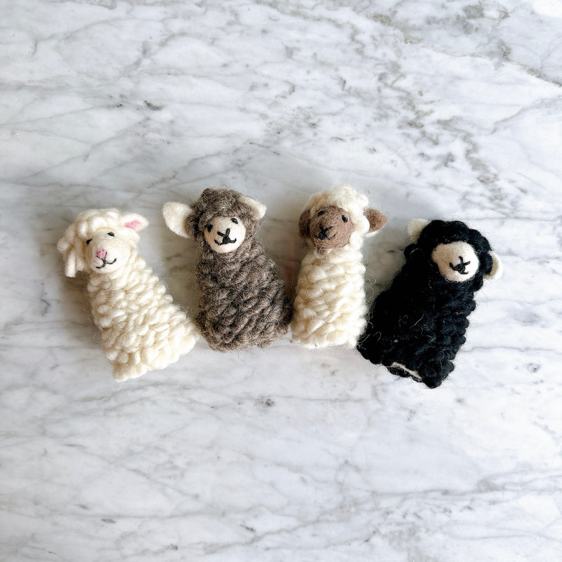 Several handmade felt sheep finger puppets are arranged in a row on a textured white marble surface. These cuddly felt sheep toys showcase a soft, fluffy texture and charming 3D designs. Visible puppets include a white sheep with pink inner ears, a brown sheep, a cream-colored sheep with a brown face, and a black sheep, each with a smiling face.