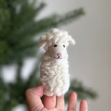 A handmade felt sheep finger puppet is held by a hand against a blurred Christmas tree and white wall. The charming white sheep puppet features a textured body resembling wool, perky ears, a pink nose, cute eyes, and a simple stitched mouth, giving it a friendly expression.
