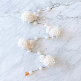 A charming wool felt sheep garland rests on a white marble surface, creating a whimsical and peaceful atmosphere. The garland features three fluffy, white sheep crafted from pure New Zealand sheep wool felt, each with a curled texture and small, light pink ears. They are evenly spaced along a thin black string. A bell dangles from one end, and a small black loop is at the top.