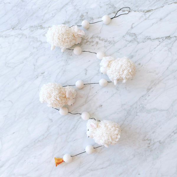 A charming wool felt sheep garland rests on a white marble surface, creating a whimsical and peaceful atmosphere. The garland features three fluffy, white sheep crafted from pure New Zealand sheep wool felt, each with a curled texture and small, light pink ears. They are evenly spaced along a thin black string. A bell dangles from one end, and a small black loop is at the top.