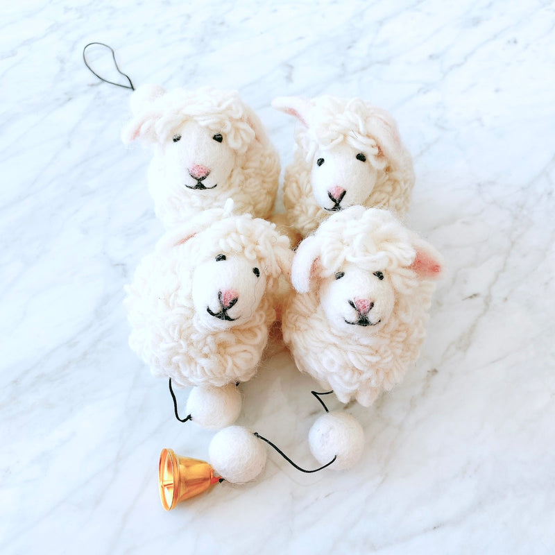 A charming felt sheep garland is displayed on a white marble surface, creating a cheerful presentation. It features three plush, off-white wool felt sheep, each with sweet pink accents on its nose and inner ear. A thin black string connects the sheep, interspersed with round, white felt balls, and ending with a shiny copper bell. The garland's fluffy texture evokes warmth and handmade charm.