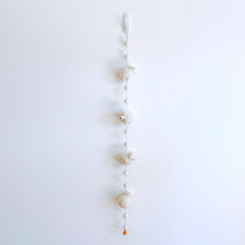 A fluffy felt sheep garland hangs against a stark white wall, featuring three evenly spaced, three-dimensional wool felt sheep. Spherical wooden beads connect the sheep, and a single copper bell hangs at the bottom, creating a serene, neutral-toned display.
