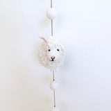 A charming wool felt sheep garland is displayed vertically against a bright white background. It features three three-dimensional white felt sheep, each with curly fleece and a pink nose, strung with white felt balls on a thin black string. A copper bell dangles whimsically at the bottom, adding a tactile charm.