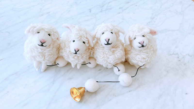 A charming wool felt sheep garland rests on a bright white marble surface. It features three fluffy white sheep, each crafted from wool felt with curly textures, small pink ears, and black stitched eyes and mouths. Black thread connects the sheep to white felt balls, leading to a copper bell at the garland's end.