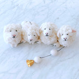 A charming sheep felt garland, featuring three white sheep and a bell, rests on a white marble-like surface, creating a calm and cheerful mood. The wool felt sheep have a fluffy texture with small pink noses and black eyes, appearing very cute. A smooth, shiny bell adds a whimsical touch at the end.