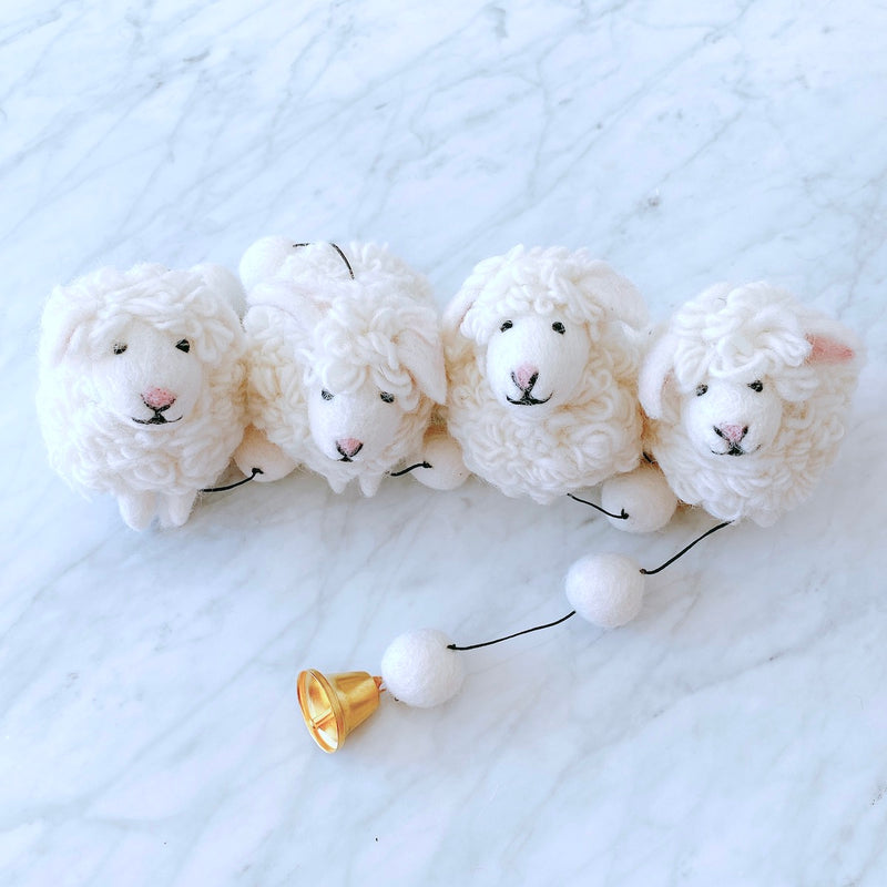 A charming sheep felt garland, featuring three white sheep and a bell, rests on a white marble-like surface, creating a calm and cheerful mood. The wool felt sheep have a fluffy texture with small pink noses and black eyes, appearing very cute. A smooth, shiny bell adds a whimsical touch at the end.