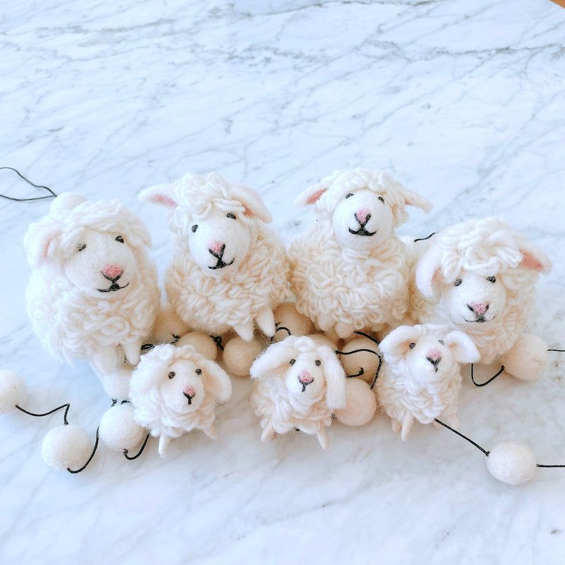 A charming wool felt sheep garland rests on a bright white marble surface. It features three fluffy off-white felt sheep, each with perky ears, a pink felt nose, and a simple stitched black mouth. The garland string connects the sheep with small, round felt balls, and a copper bell dangles from one end.