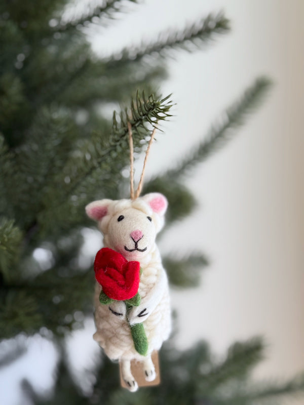 A wool felt sheep ornament holding a heart hangs on a cheerful Christmas tree. This soft, fluffy ornament features tiny pink ears, a pink stitched nose, and closed stitched eyes, with a jute string for hanging.