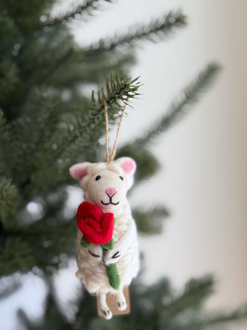 A wool felt sheep ornament holding a heart hangs on a cheerful Christmas tree. This soft, fluffy ornament features tiny pink ears, a pink stitched nose, and closed stitched eyes, with a jute string for hanging.