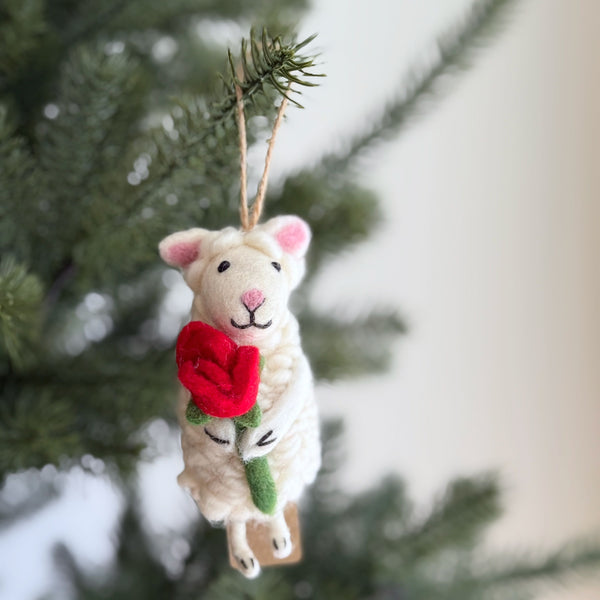 A cream-colored wool felt sheep ornament hangs on a festive Christmas tree, evoking warmth and nostalgia. This woolly, soft sheep features pointy pink ears, a cheerful smile, and holds a red rose with a green stem, adorned with a needle-felted heart. A jute string is attached for easy display.