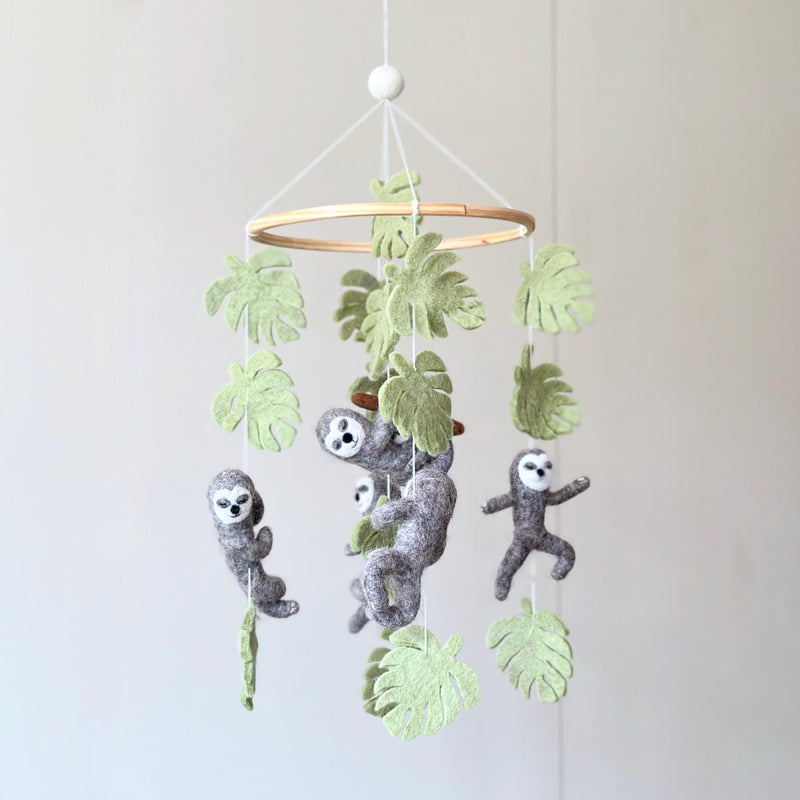 A Wool Felt Sloth Baby Mobile hangs suspended against a plain, light gray wall, creating a whimsical and calming scene. This charming nursery baby mobile features a light-colored wooden frame from which three gray wool felt sloths dangle by thin white cotton threads. The sloths are in various adorable poses, with bendable limbs adding to their playful design, ideal for a baby's room.