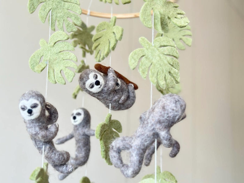 A close-up shot shows a charming wool felt sloth baby mobile with a neutral background. Four adorable gray wool felt sloth figures, each in a unique pose with detailed facial features, hang from a wooden frame by white cotton threads. The mobile's soft textures and colors are emphasized.