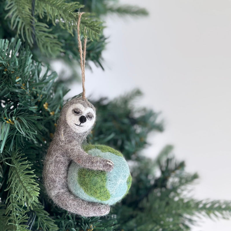 A handcrafted felt sloth ornament is showcased on a lush green Christmas tree. This unique Christmas ornament features a gray sloth with a light gray face, closed eyes, and a smiling mouth, gently embracing a blue and green globe. It evokes a whimsical sense of environmental awareness, perfect as an Earth-holding decor piece.